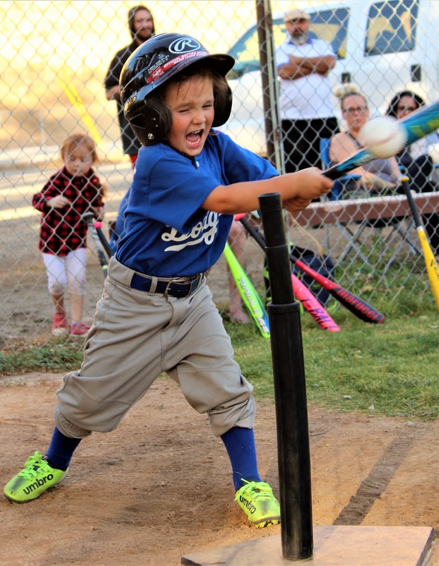 A small child (perhaps about five years old) enthusiastically hits a baseball off of a tee at a tee ball game.