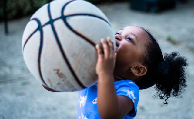 A young child (1 or 2 years old, perhaps), smiles as she hoists a basketball up in front of her face as she gazes over it. She appears to be about to "shoot" the ball toward some hoop that is out of view.