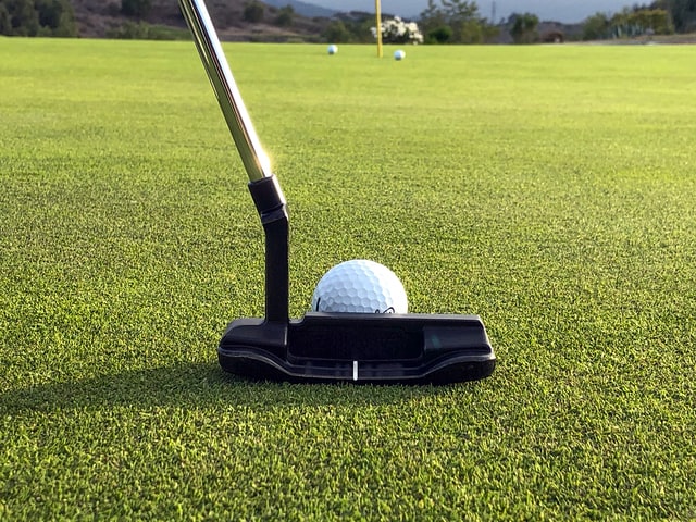 A close-to-ground shot of a golf putter about to hit a golf ball toward a flag (in a hole) in the distance, flanked by two previously-putted golf balls.